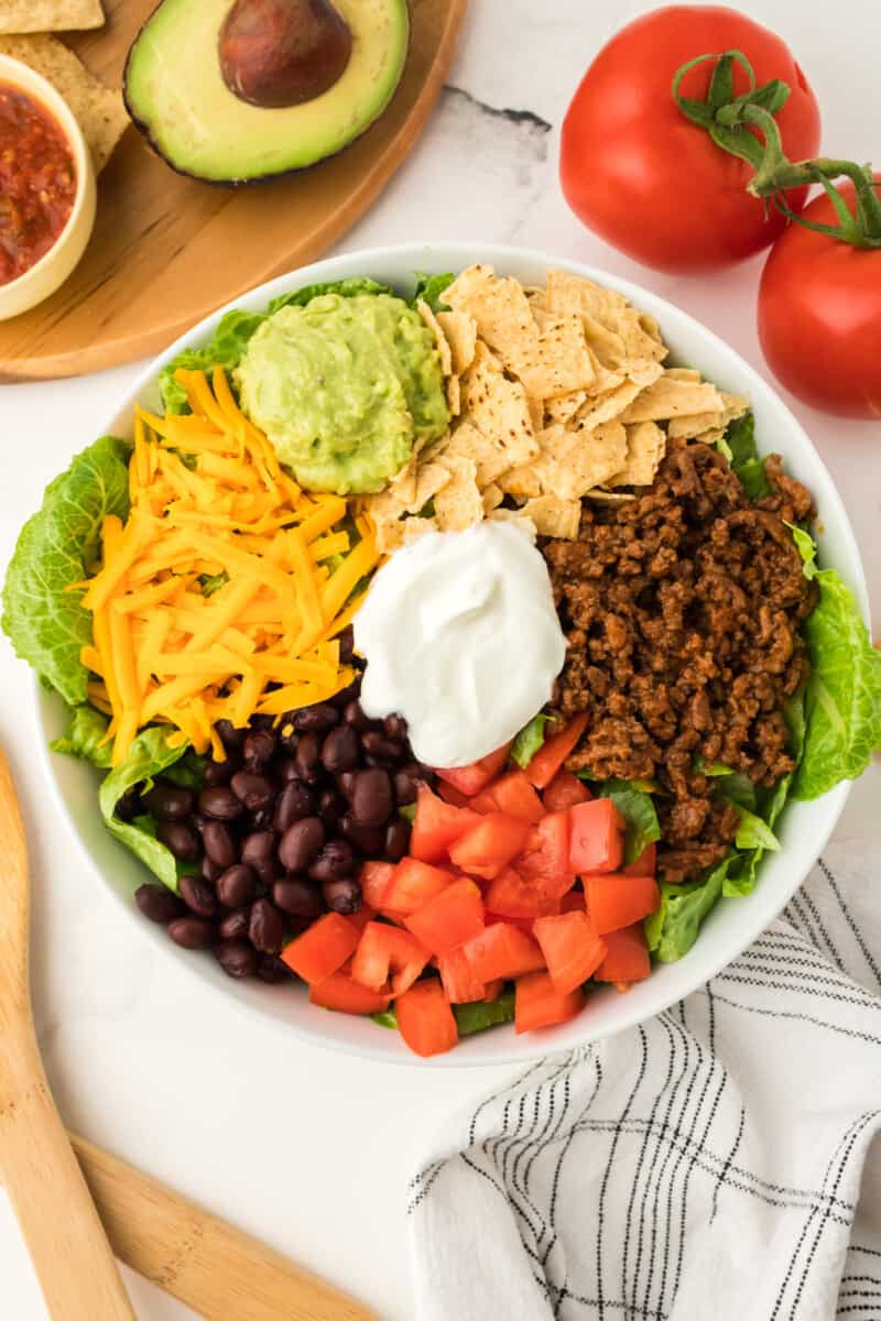 a bowl of greens filled with ground beef, tomatoes, black beans, shredded cheese tortillas strips, guacamole, and sour cream