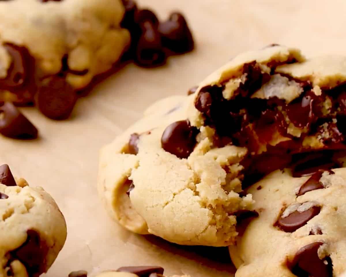 Chocolate chip cookie broken open to reveal a soft, gooey center with melted chocolate, surrounded by more golden cookies on parchment paper