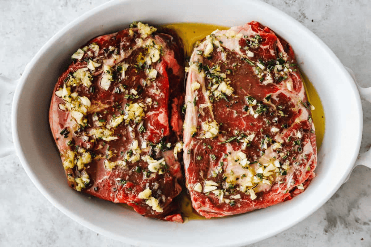 Two ribeye steaks in a shallow white dish covered in marinade.