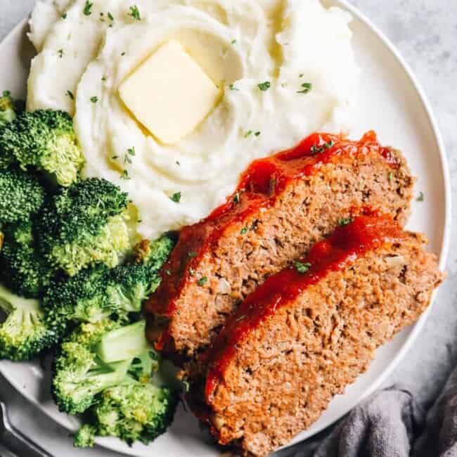 overhead view of 2 slices of crockpot meatloaf on a white plate with broccoli and mashed potaotes.