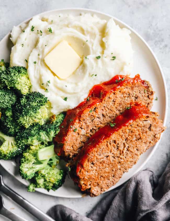 overhead view of 2 slices of crockpot meatloaf on a white plate with broccoli and mashed potaotes.