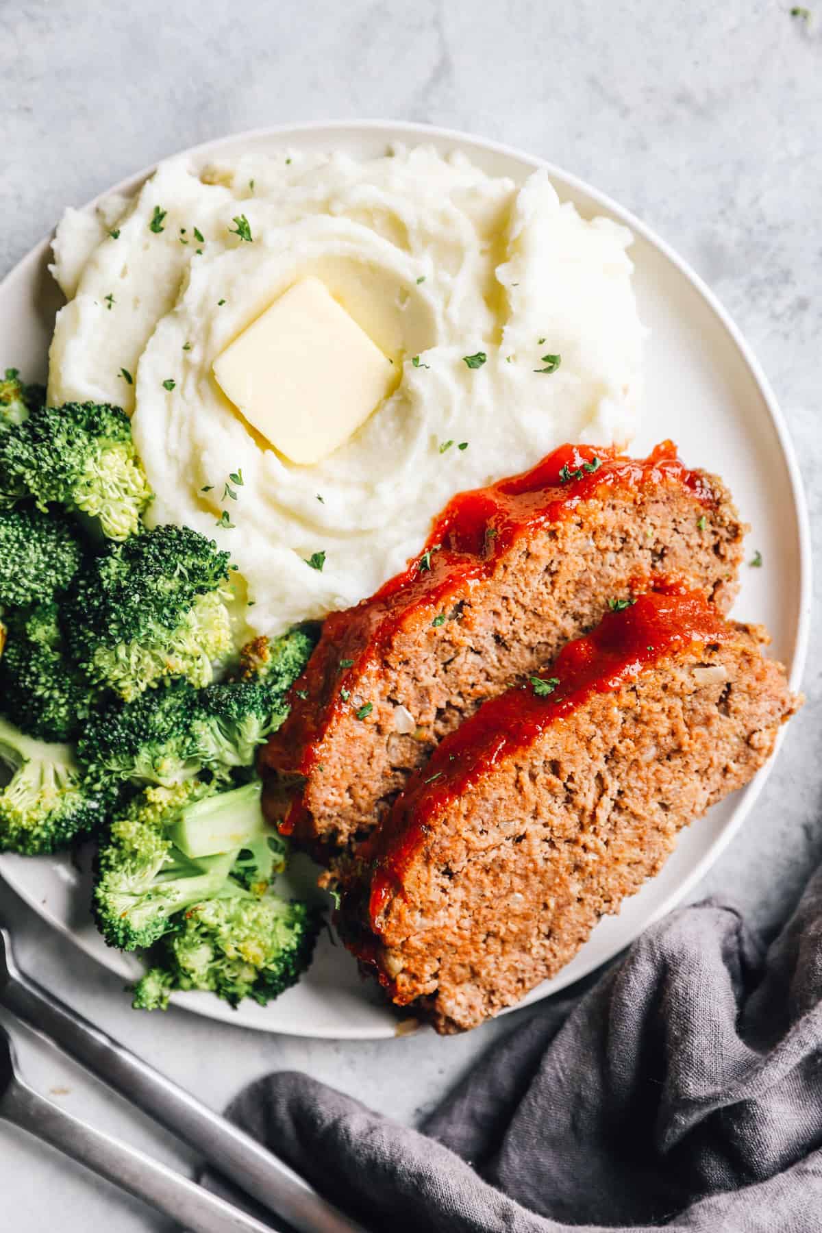 Overhead view of 2 slices of meatloaf on a white plate with broccoli and mashed potaotes.