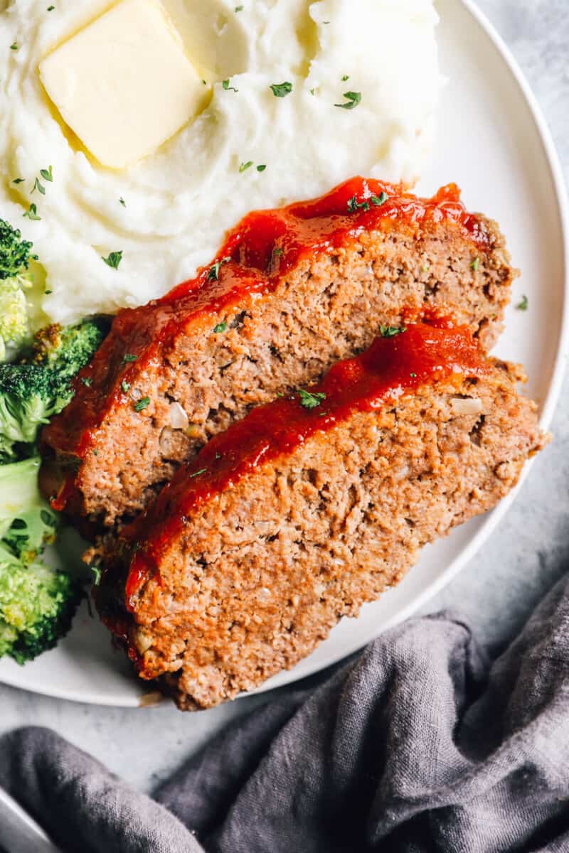 Two slices of meatloaf on a white plate with broccoli and mashed potatoes.