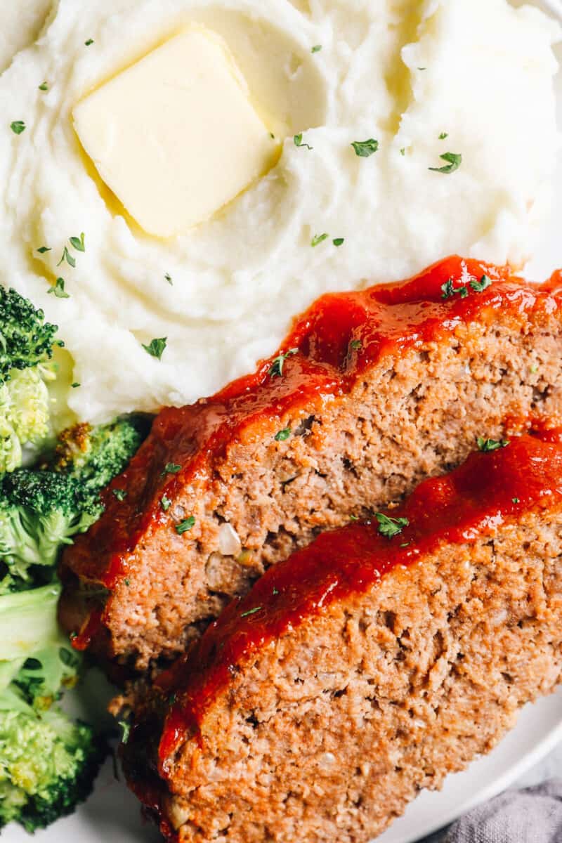 2 slices of crockpot meatloaf on a white plate with broccoli and mashed potaotes.