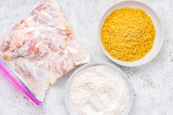 overhead view of air raw chicken pieces marinating in buttermilk in a ziplock bag next to flour and cornflakes in glass bowls.