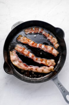 4 strips of bacon frying in a cast iron skillet.