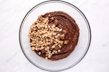 overhead view of chopped peanut butter cups and peanut butter baking chips added to peanut butter brownie batter in a glass bowl.