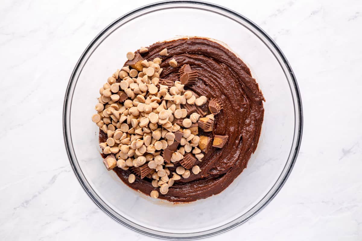Overhead view of chopped peanut butter cups and peanut butter baking chips added to peanut butter brownie batter in a glass bowl.