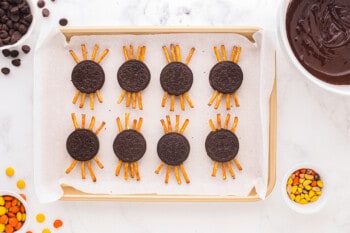 oreos lined up on a baking tray with pretzel sticks attached like legs