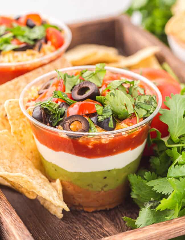 a 7 layer dip cup on a wooden tray with tortilla chips.