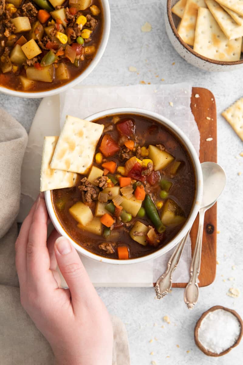 overhead view of a hand grabbing a bowl of hamburger soup with saltine crackers.