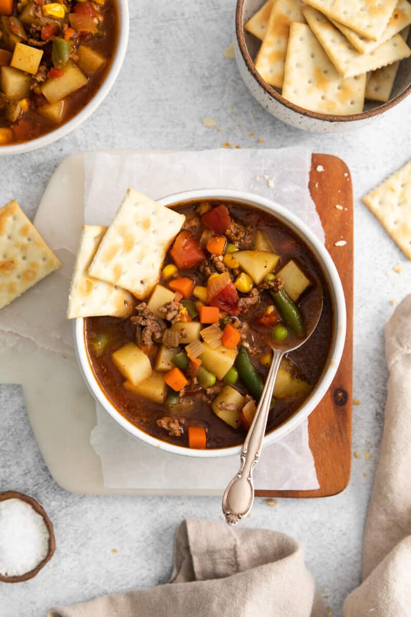 overhead view of hamburger soup in a white bowl with saltine crackers and a spoon.