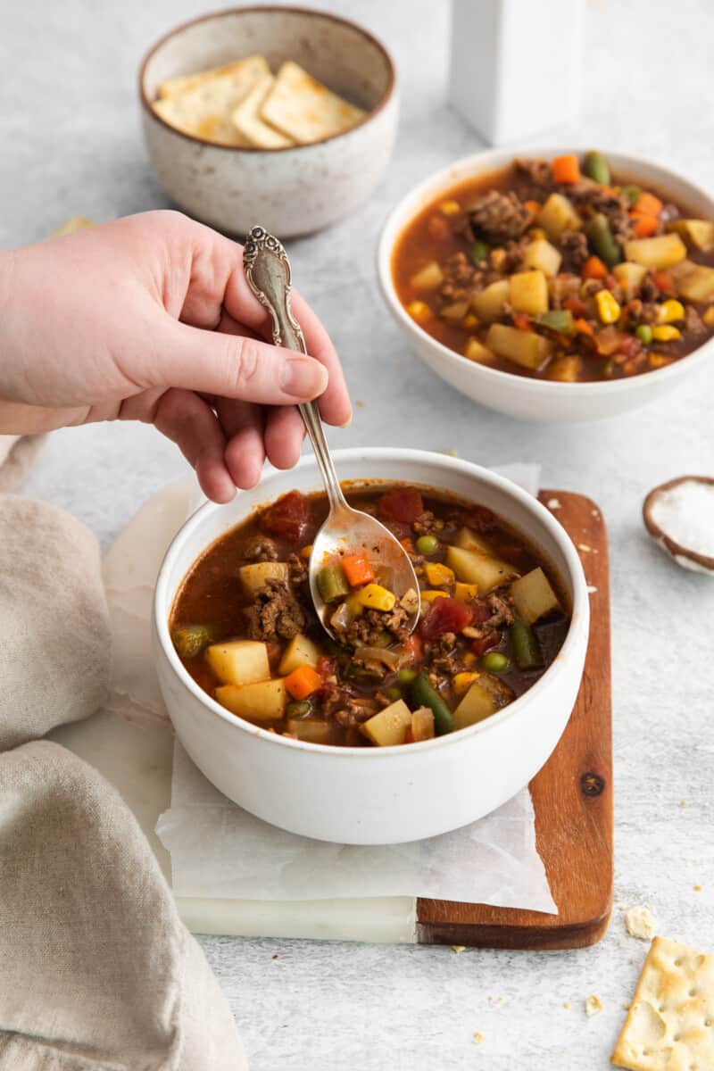 a hand using a spoon to scoop a spoonful of hamburger soup from a white bowl.