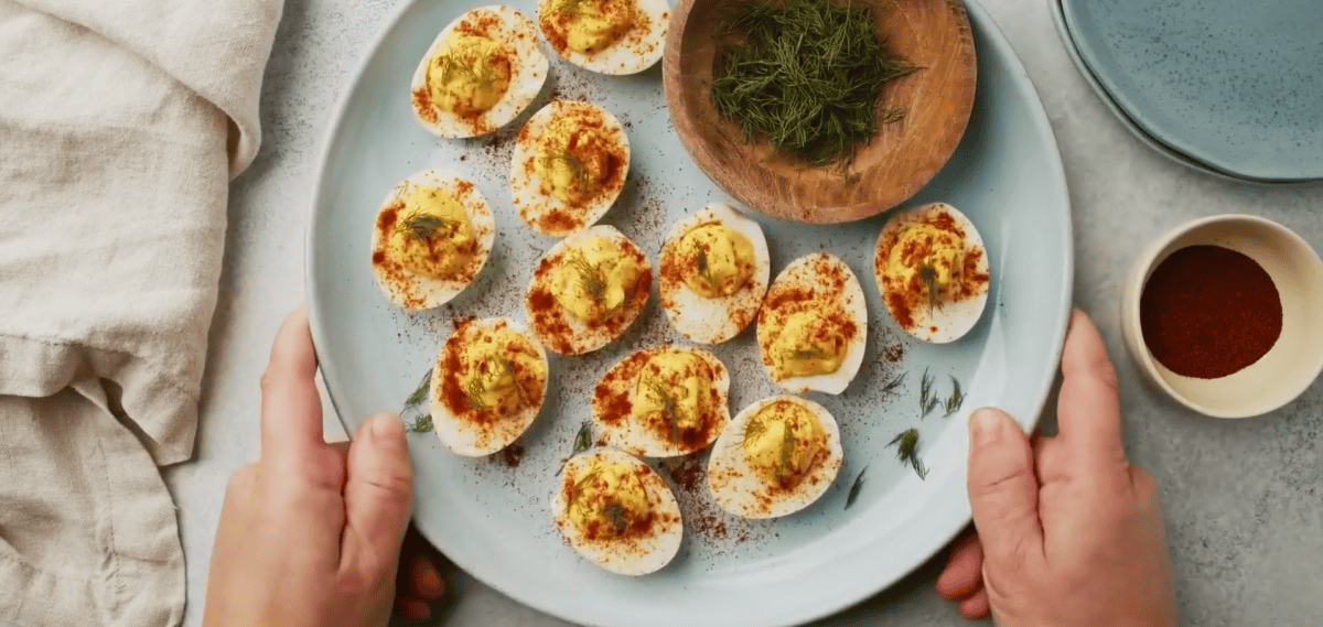 deviled eggs garnished with smoked paprika and fresh dill on a white serving platter with a bowl of fresh dill and a bowl of smoked paprika.