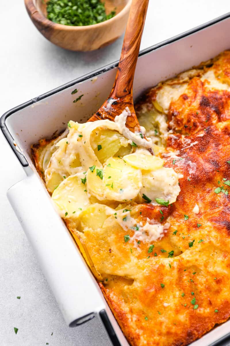 a wooden spoon scooping au gratin potatoes out of a 9x13 baking pan.