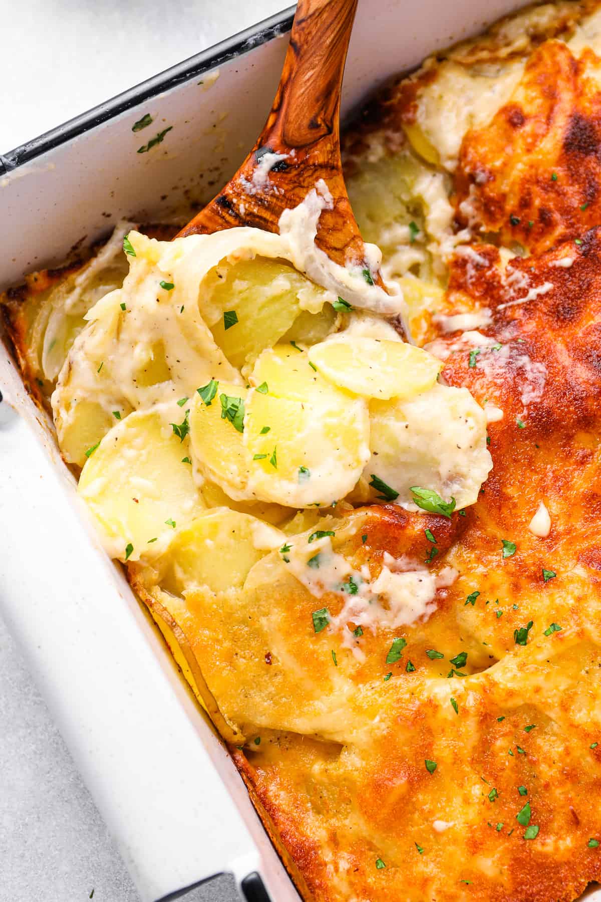 a wooden spoon scooping au gratin potatoes out of a 9x13 baking pan.