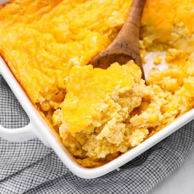 a wooden spoon scooping corn casserole from a white baking dish.