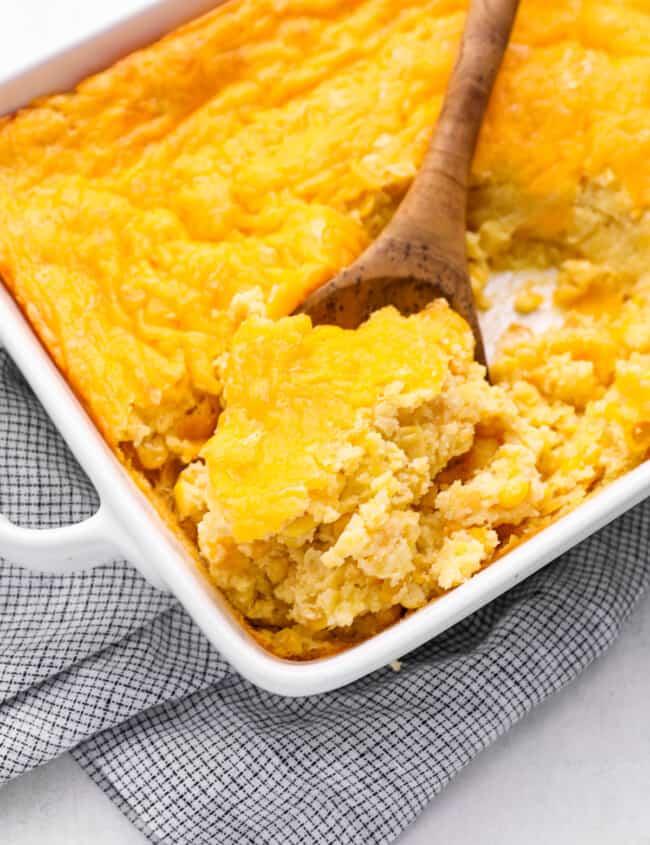 a wooden spoon scooping corn casserole from a white baking dish.