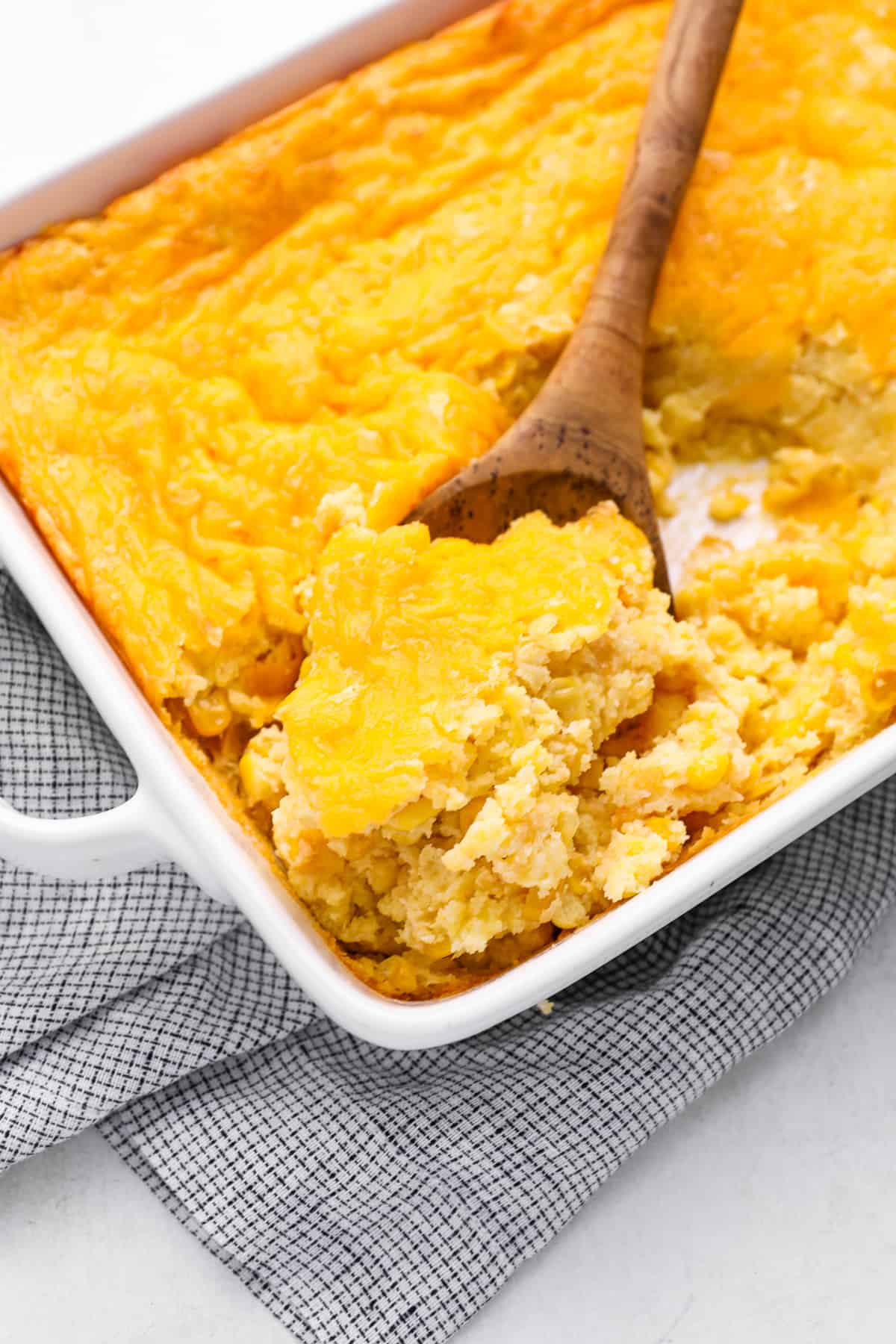 a wooden spoon scooping corn casserole from a white baking dish.