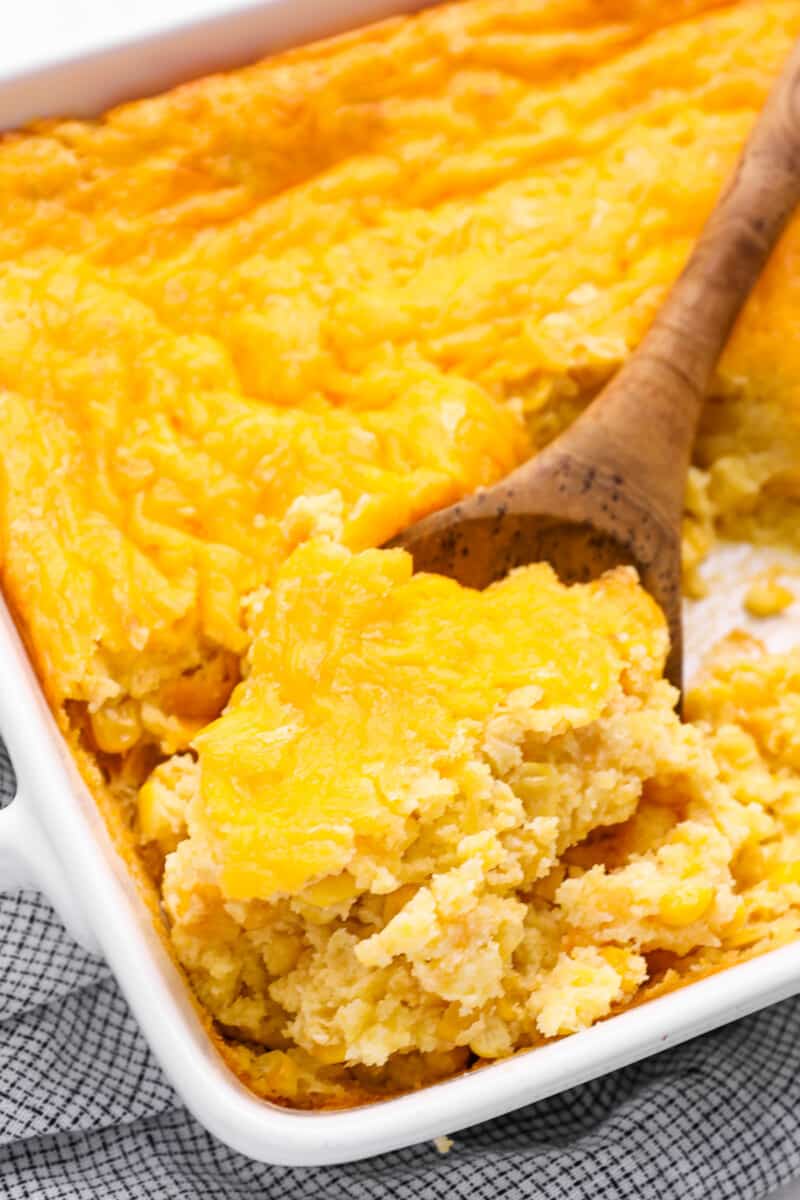 close up of a wooden spoon scooping corn casserole from a white baking dish.