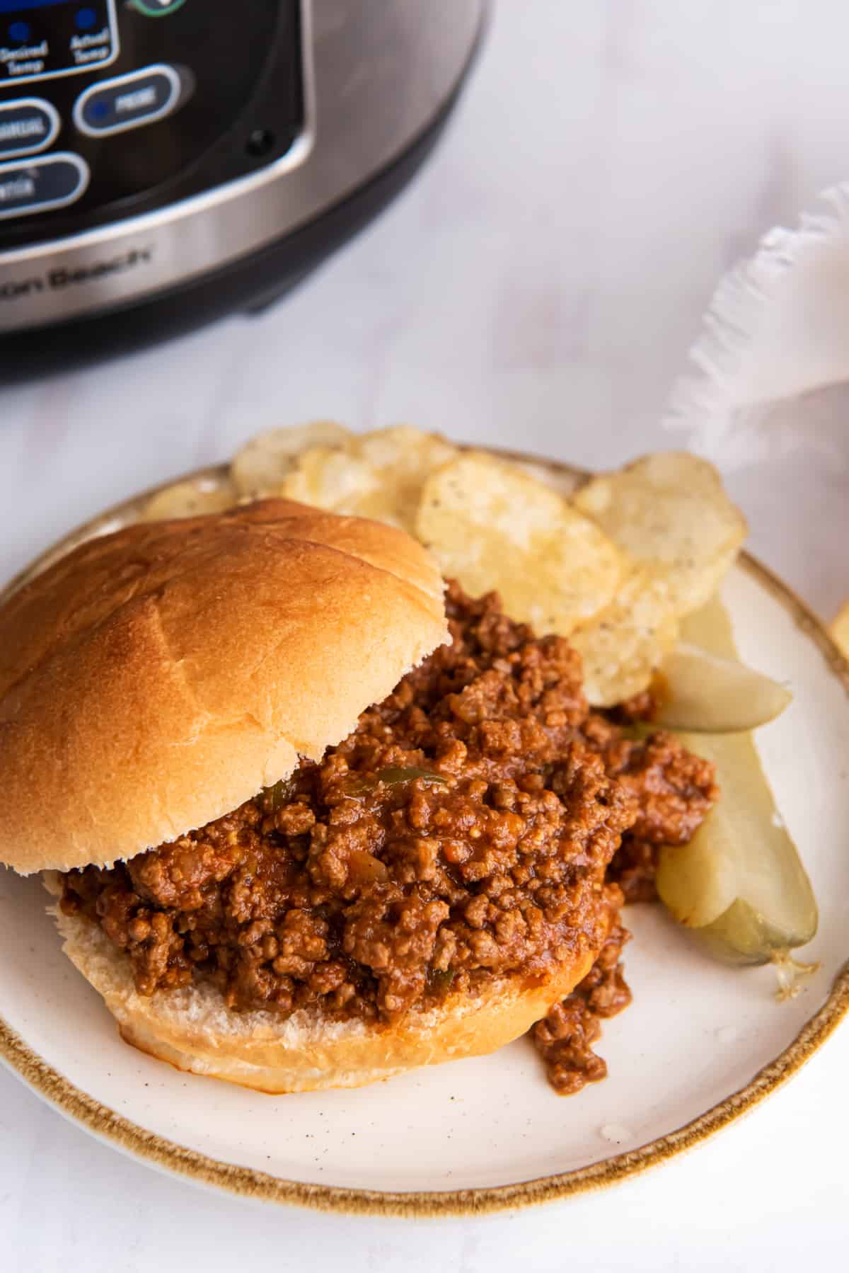 slow cooker sloppy joe meat on a hamburger bun on a plate with chips and a pickle.