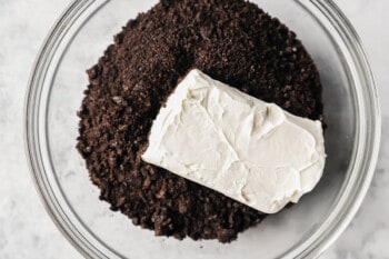 a brick of cream cheese added to oreo crumbs in a glass bowl.