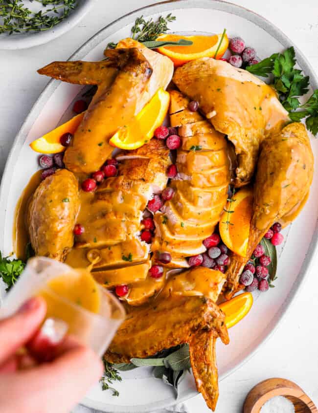 a hand pouring turkey gravy over a sliced whole turkey on a white plate.