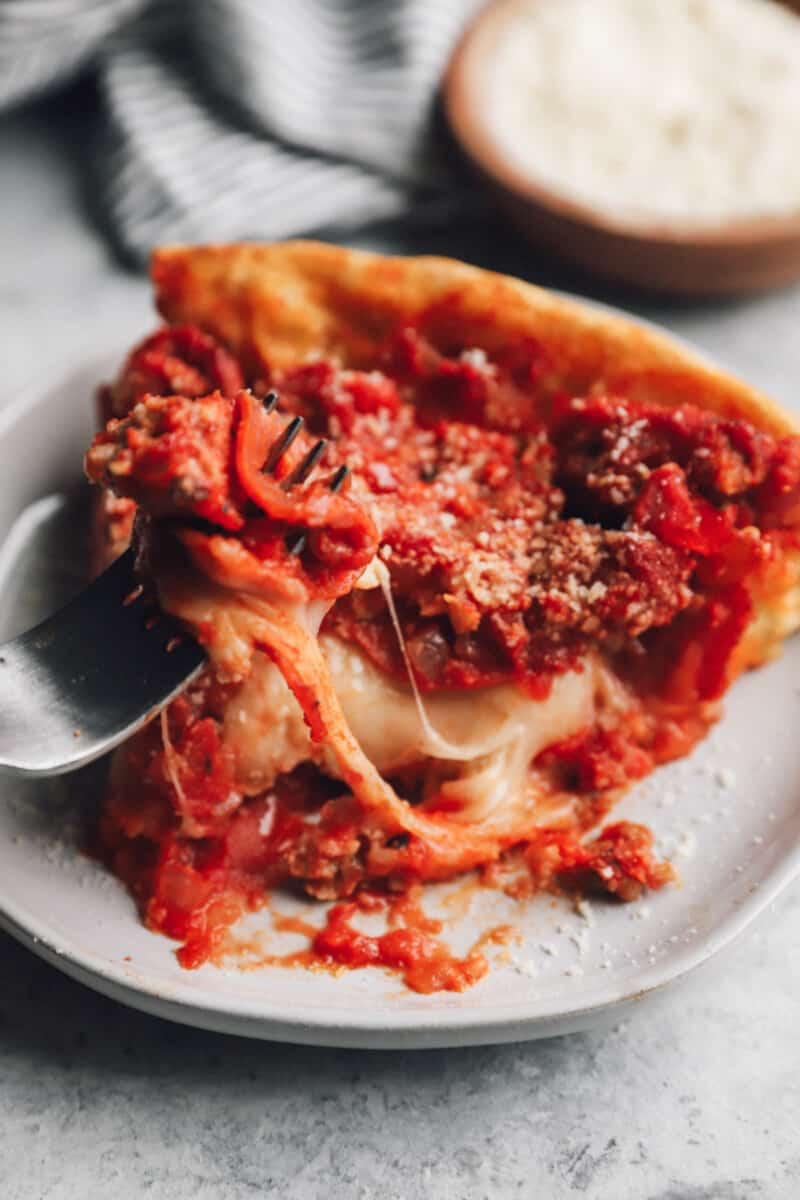 a fork removing a bite of chicago deep dish pizza from a slice on a white plate.