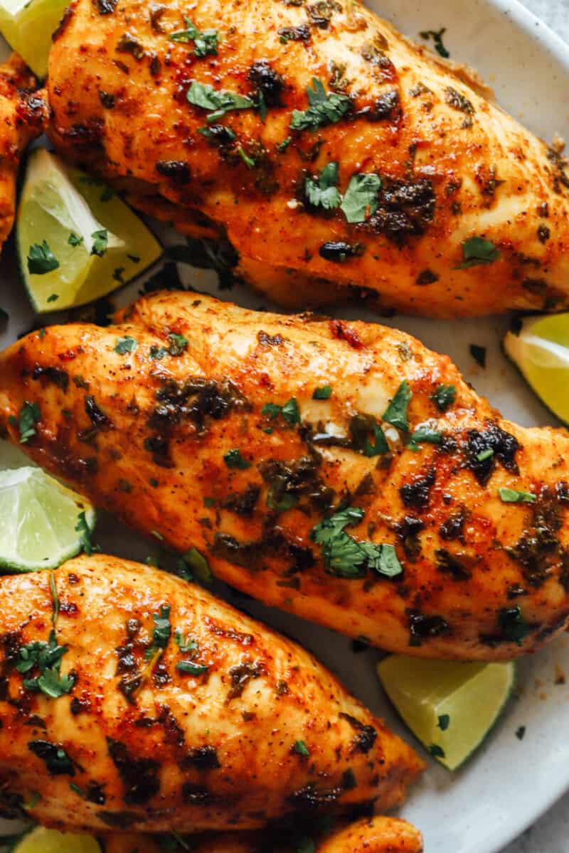 close up of 3 cilantro lime chicken breasts surrounded by lime wedges on a white plate.