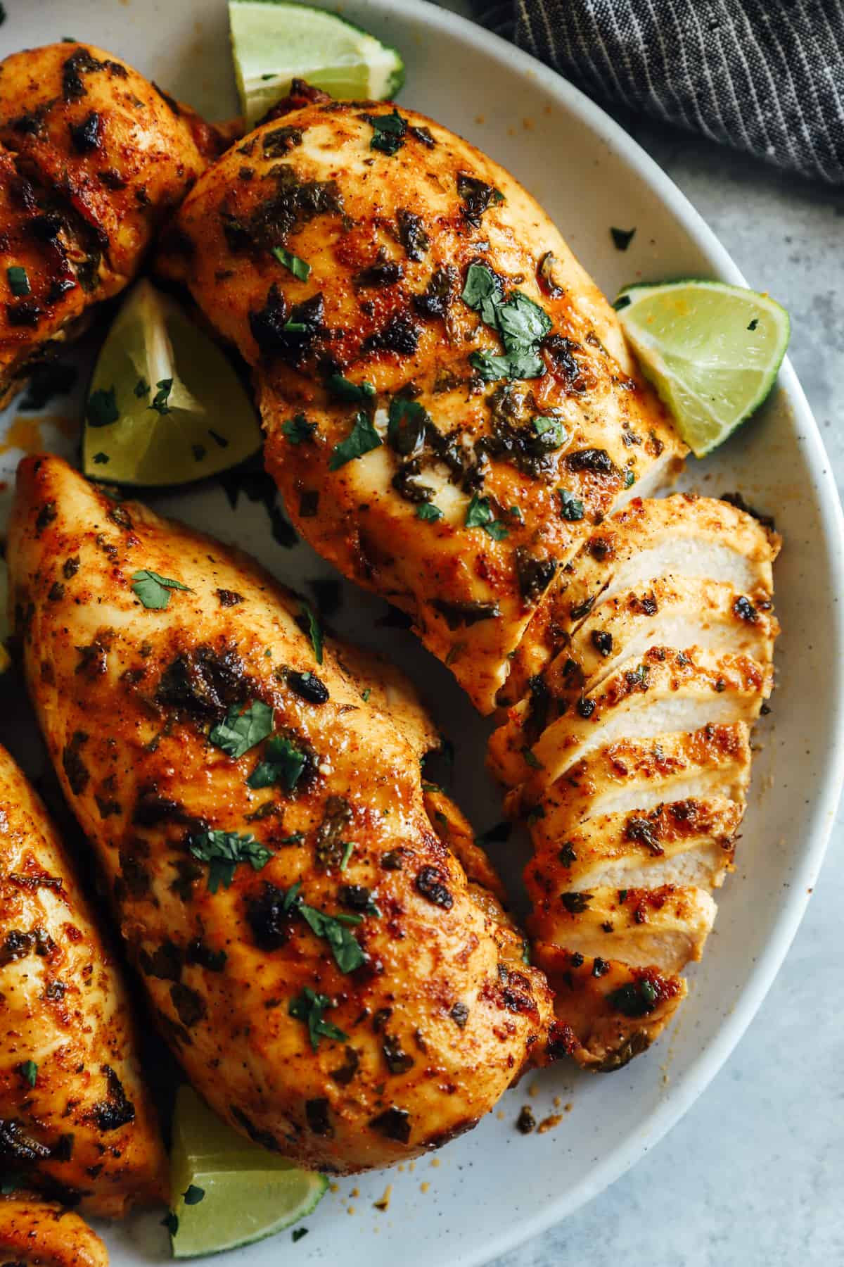 a partially sliced cilantro lime chicken breast surrounded by more chicken breasts and lime wedges on a white plate.