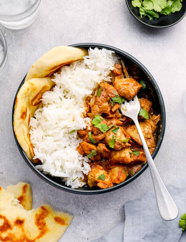 overhead view of crockpot chicken tikka masala with rice and naan in a black bowl with a fork.