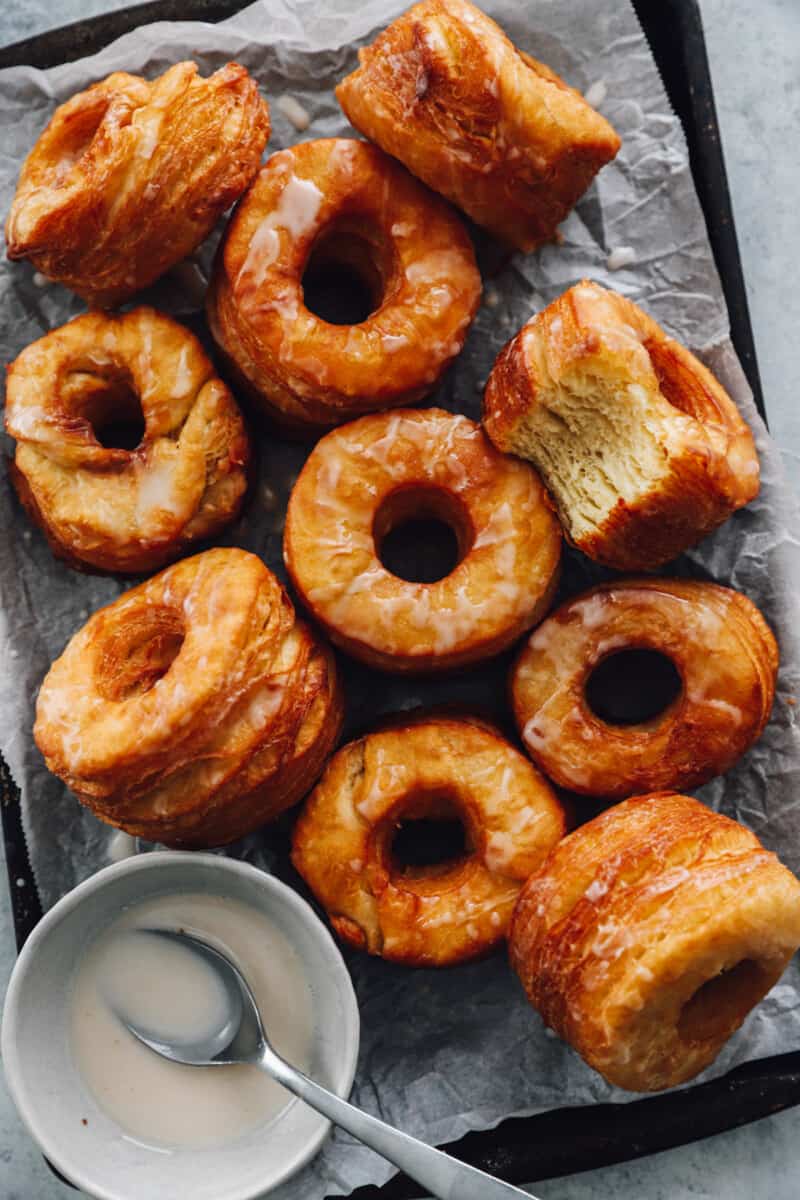 a bitten cronut next to 9 cronuts on a baking sheet with glaze in a white bowl with a spoon.