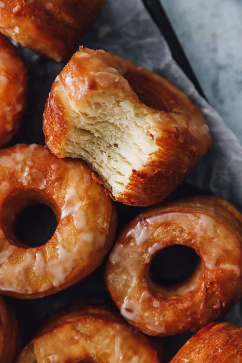 close up of a bitten cronut next to more cronuts on a baking sheet.