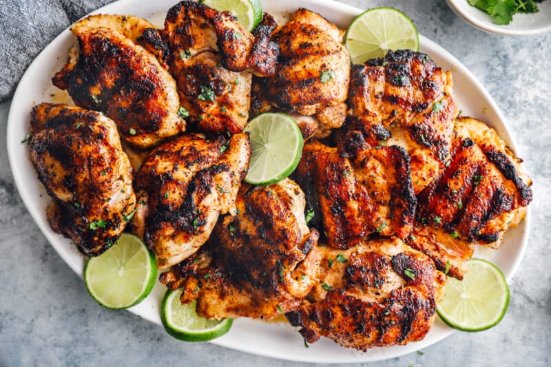 overhead view of grilled chicken thighs on a white oval serving platter with lime slices.