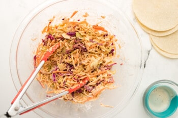 coleslaw in a glass bowl with orange tongs.