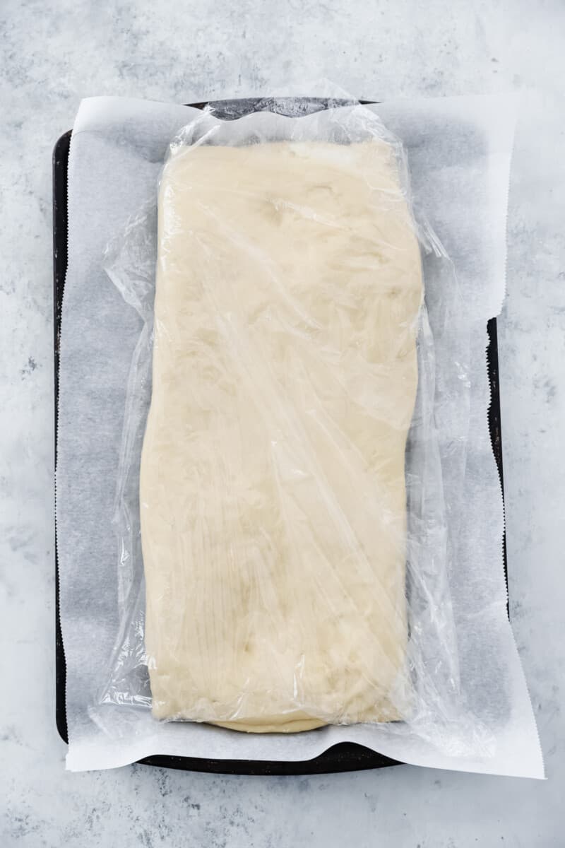 Folded cronut dough wrapped in plastic wrap on a lined baking sheet.