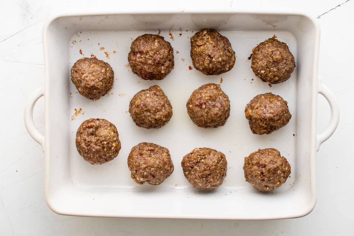 11 partially cooked meatballs in a white baking pan.