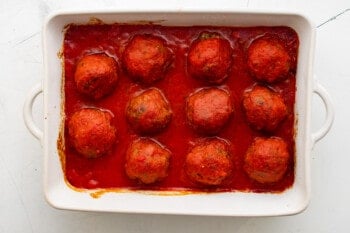 sauce poured over 11 meatballs in a white baking pan.