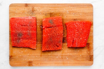 3 salmon filets seasoned with salt and pepper on a wooden cutting board.
