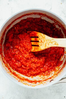overhead view of new york style pizza sauce in a white bowl with a slotted wooden spoon.