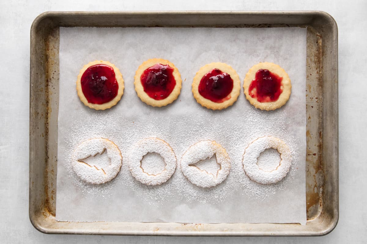 Cookie on a tray, four are topped with jam, the others have shapes cut out and are topped with powdered sugar.