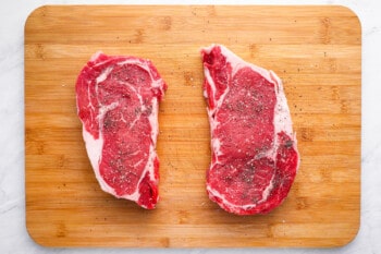 2 seasoned raw ribeye steaks on a cutting board.
