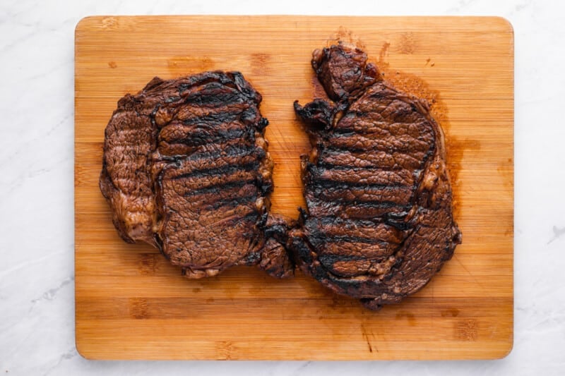 2 grilled ribeyes on a wooden cutting board.