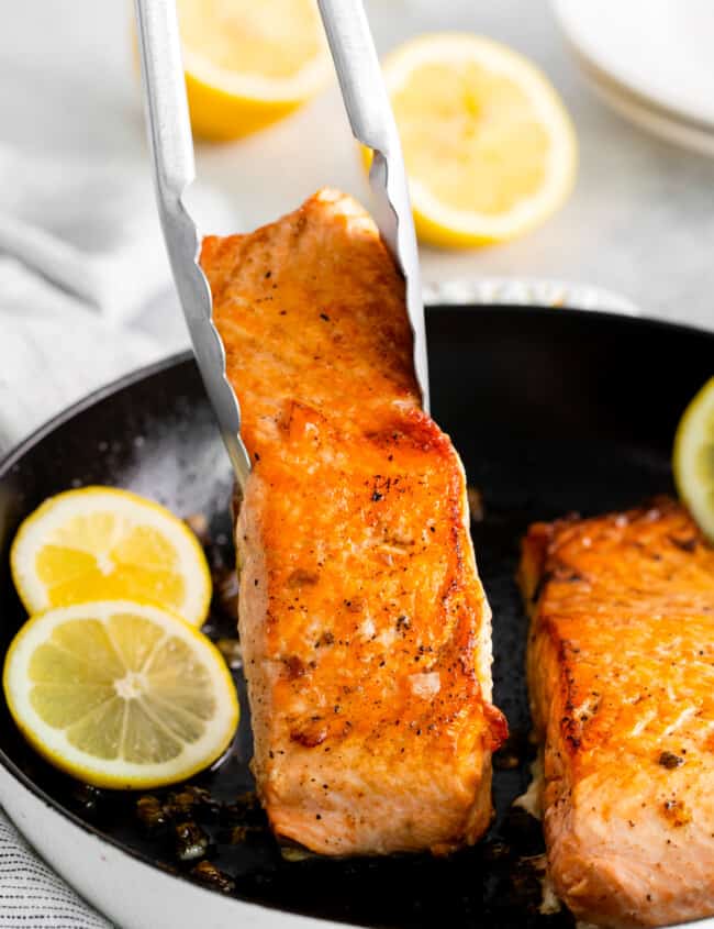 tongs grabbing a salmon filet from a cast iron pan.