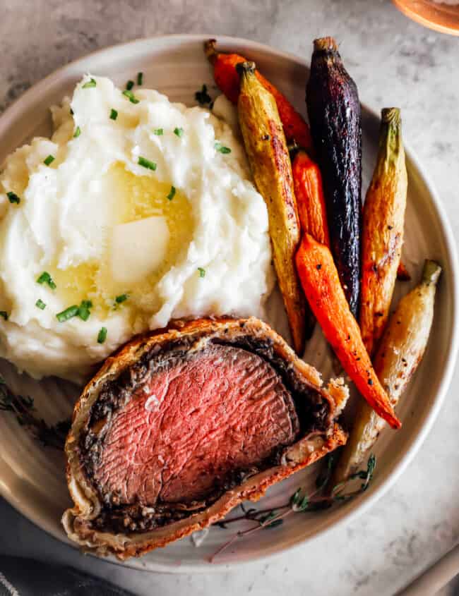 a slice of beef wellington on a white plate with carrots and mashed potatoes.