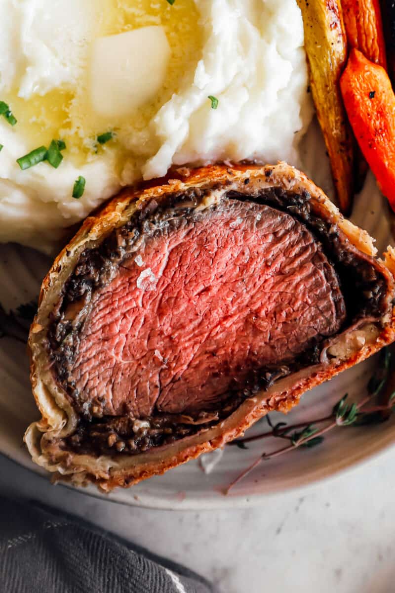close up of a slice of beef wellington on a white plate with carrots and mashed potatoes.