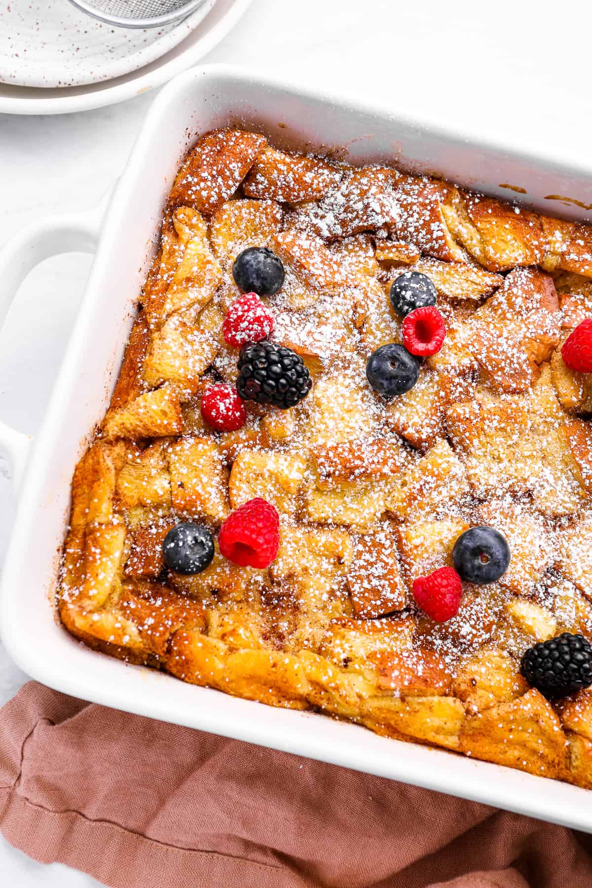 bread pudding in a white baking pan with powdered sugar and berries.