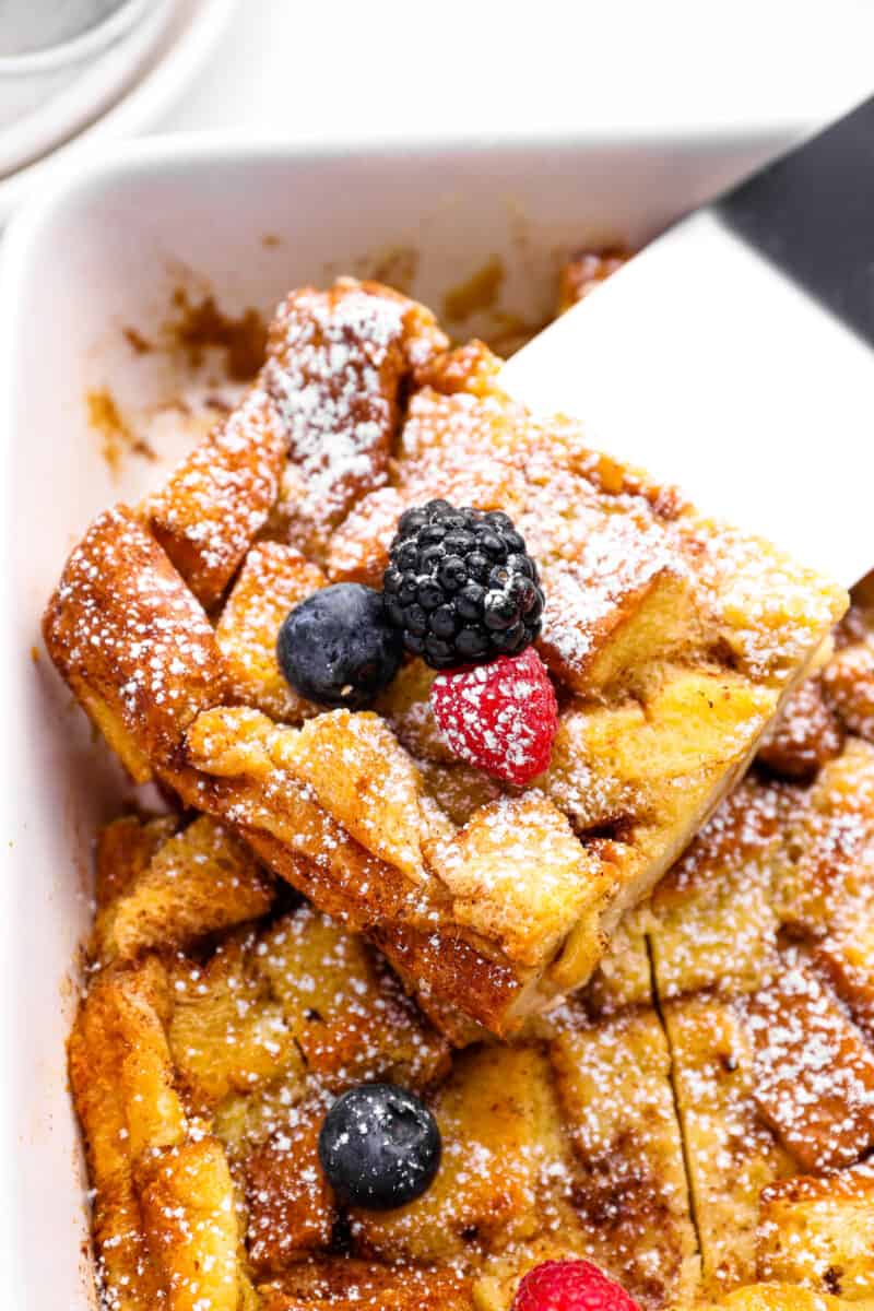 close up of a slice of bread pudding being lifted out of a white baking pan with a ribber spatula.