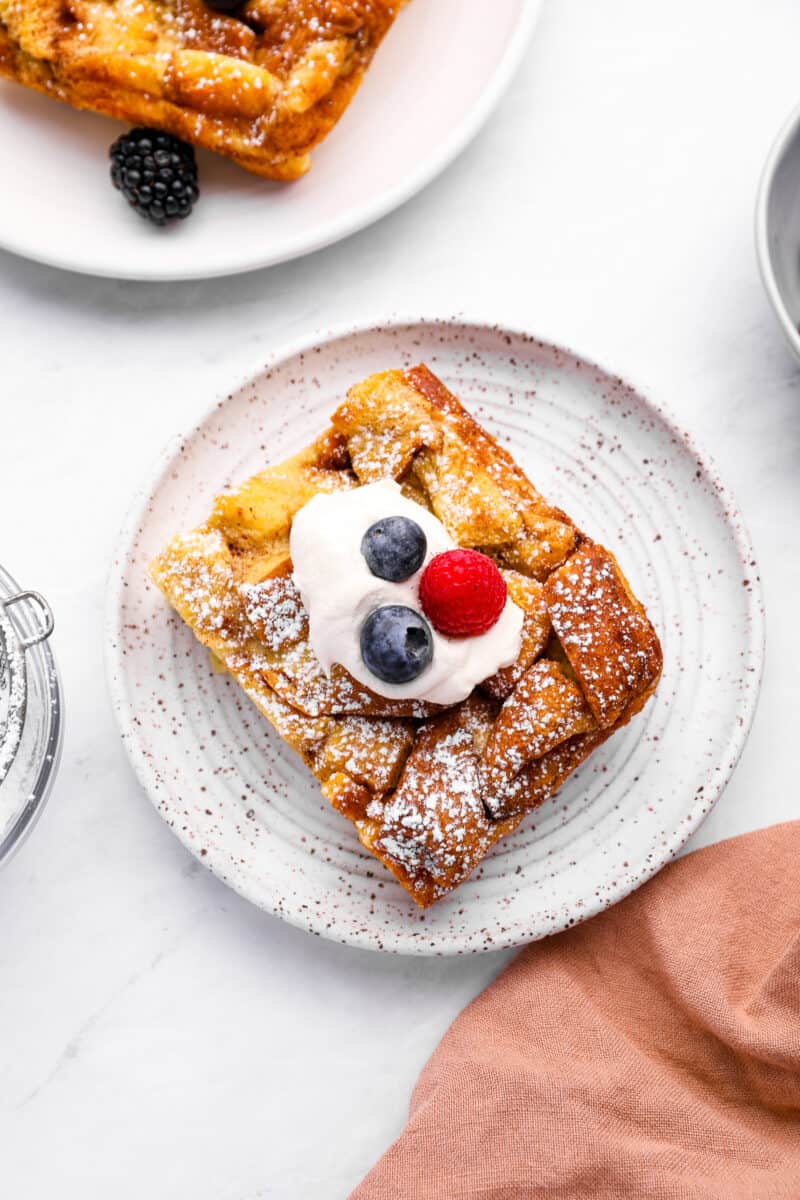 a serving of bread pudding on a white plate with berries and whipped cream.