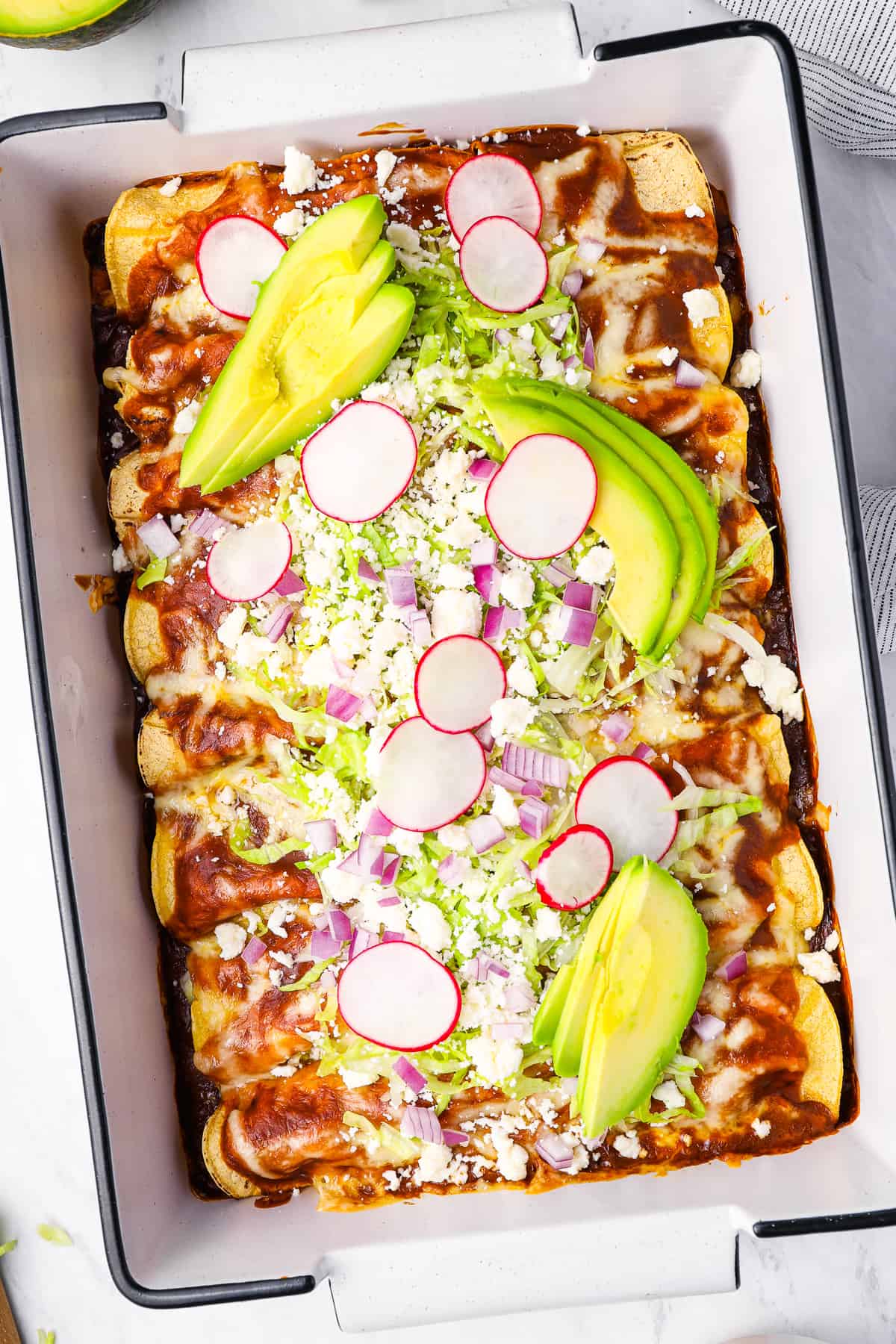overhead view of 8 baked chicken enchiladas topped with enchilada sauce, cheese, lettuce, radish, onion, and avocado.